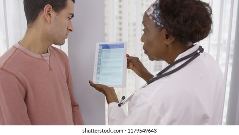 Senior African MD Showing Male Latino Patient Different Health Plans On Tablet