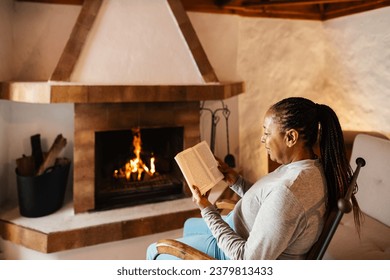 Senior African woman reading book while warming in front of fireplace in her house - Powered by Shutterstock