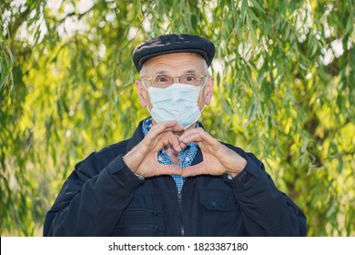 Senior Aged Man Wearing Medical Blue Facemask Glasses And Cap Showing Heart Gesture Outside