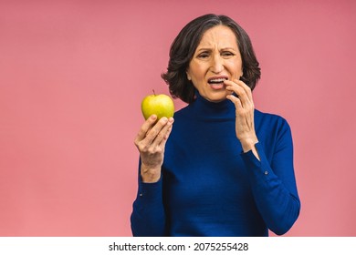 Senior Aged Mature Woman Touching Mouth With Hand With Painful Expression Because Of Toothache Or Dental Illness On Teeth Holding Green Apple. Dentist Concept Isolated Over Pink Background.