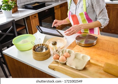 Senior Aged Woman Baking In Home Kitchen. Using Tablet Computer To Find Recipe. Mature Woman Cooking With The Help Of Internet. Modern Technology For Older People.