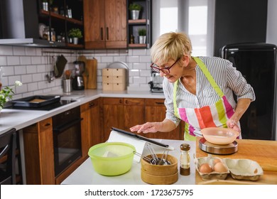 Senior Aged Woman Baking In Home Kitchen. Using Tablet Computer To Find Recipe. Mature Woman Cooking With The Help Of Internet. Modern Technology For Older People.