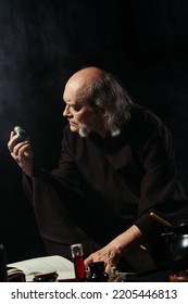 Senior Alchemist Holding Vial With Magic Ingredient Near Flasks And Magic Cookbook Isolated On Black