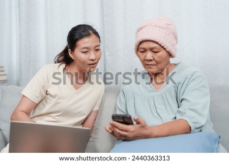 Senior in aqua tunic and knit cap attentively examines smartphone, guided by young woman with laptop. Concentration and curiosity blend as aged female in teal dress learns about phone from younger