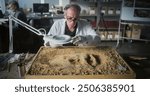 Senior archaeologist cleans ancient extinct human or animal bones in sand after excavation using brush and tools, studies fossil remains in archaeological lab. Male colleague works in the background.