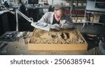 Senior archaeologist cleans prehistoric human or animal bones after excavation using tools and brush, examines ancient fossil remains in archaeological laboratory. Colleague works in the background.