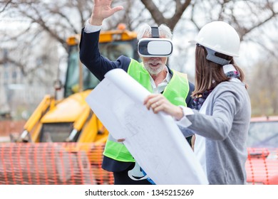 Senior Architect Or Businessman Using Virtual Reality Goggles On A Construction Site. Young Female Colleague Holding Building Blueprints And Explaining Construction Details.