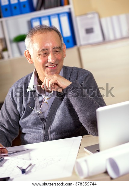 Senior architect working on\
construction blueprint in office, he is looking at\
laptop.