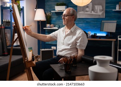 Senior art teacher sketching feeling inspired sketching vase on paper canvas using sharp pencil in home studio. Portrait of retired talented elederly man doing creative masterpiece on easel.