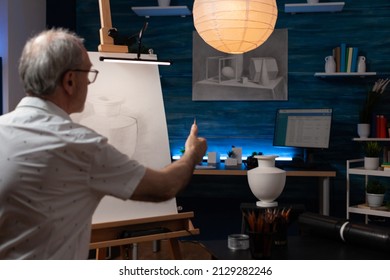 Senior artist feeling inspired looking at plaster vase using sharp pencil for scaling doing creative pencil drawing. Elderly man in the evening sketching still life on easel in home art studio. - Powered by Shutterstock