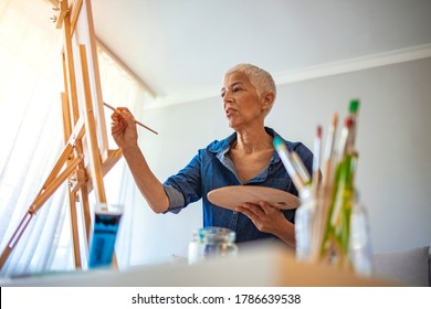 Senior artist working in concentration. Side view portrait of white haired senior woman holding palette painting pictures at easel in art studio standing against windows in sunlight - Powered by Shutterstock