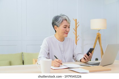 Senior Asia Woman Working From Home Using Laptop And Checking Message On Smart Phone.