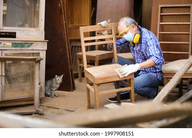 Senior Asian carpenter man is sanding wooden chair in his own garage style workshop at retirement home while his cat is watching for hobby with copy space - Shutterstock ID 1997908151