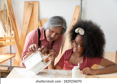 Senior Asian Carpenter Teaching A Girl Painted The Wooden Bird Nest. A Little African American Inventor Practice To Be A Carpenter In Carpentry Shop. DIY Woodworking Crafts And Hobbies Concepts