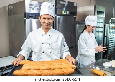 A senior Asian chef male  bakers in a white chef dresses uniform and hats standing cutting on a counter with many baked and unbaked bread on trays at a bakery kitchen restaurant. - Powered by Shutterstock