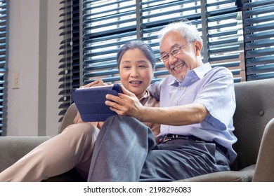 Senior Asian Couple In Retirement Age Looking At Nostalgic Photo Using Digital Tablet While Sitting On The Sofa Couch In Their Retirement Home For Elder And Pension Activities