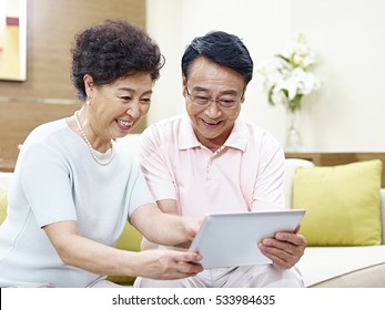 Senior Asian Couple Sitting On Couch Looking At Tablet Computer Together, Happy And Smiling