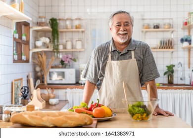 Senior Asian Elder Man Happy Living In Home Kitchen. Grandfather Cooking Salad Dish Standing At Counter With Happiness And Smile Enjoy Retirement Life. Older People Activity In Family At Home.