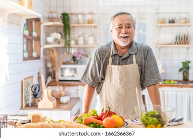 Senior Asian Elder Man Happy Living In Home Kitchen. Grandfather Cooking Salad Dish Standing At Counter With Happiness And Smile Enjoy Retirement Life. Older People Activity In Family At Home.