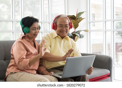 Senior Asian Elderly Couple In Home Casual Outfit With Happy Smiling Emotion Sitting In Living Room Wearing Headphone And Using Laptop To Video Call Facetime With Family Together