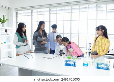 Senior Asian female teacher or scientist teaching group of diverse students in laboratory. young children using tablets and laptops, listening attentively. chemical test tubes and microscope in lab - Powered by Shutterstock