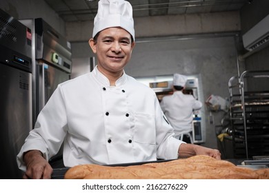 Senior Asian Male Chef In White Cook Uniform And Hat Showing Tray Of Fresh Tasty Bread With A Smile, Looking At Camera, Happy With His Baked Food Products, Professional Job At Stainless Steel Kitchen.