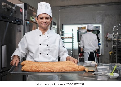 Senior Asian male chef in white cook uniform and hat showing tray of fresh tasty bread with a smile, looking at camera, happy with his baked food products, professional job at stainless steel kitchen. - Powered by Shutterstock