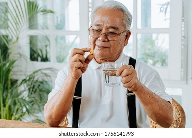 Senior Asian Man With Cup Of Water And Pill Taking Remedy In Cozy Room At Home. Old Male Taking Medicine