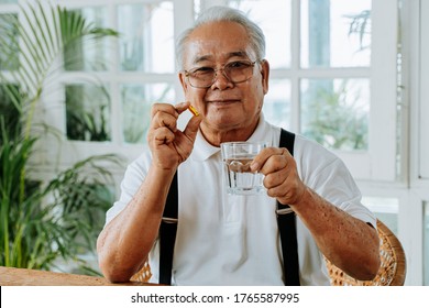 Senior Asian Man With Cup Of Water And Pill Looking At Camera And Taking Remedy In Cozy Room At Home. Old Male Taking Medicine.