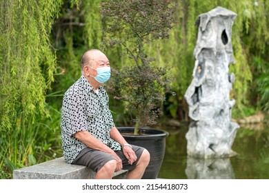 Senior Asian Man With Face Mask On, Sitting On A Bench. Outdoor Garden Setting.