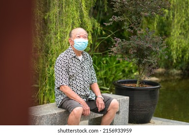 Senior Asian Man With Face Mask On, Sitting On A Bench. Outdoor Garden Setting.