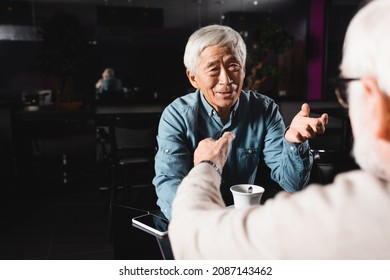 Senior Asian Man And His Blurred Friend Gesturing During Conversation In Cafe