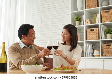 Senior Asian Man And His Young Daughter Celebrating Fathers Day Together: They Holding Glasses Of Wine In Hands And Clinking Them Together