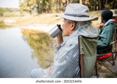 Senior Asian Man Holding Thermo Cup Near Blurred Friends In Fishing Outfit Near Lake