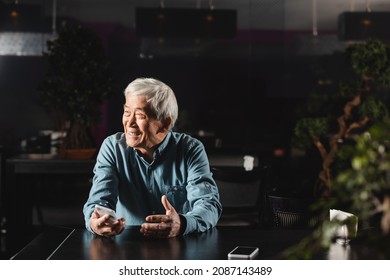 Senior Asian Man Looking Away And Smiling While Sitting With Mobile Phone In Cafe