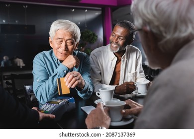 Senior Asian Man Paying Near Credit Card Reader In Cafe Near Multiethnic Friends