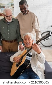 Senior Asian Man Playing Acoustic Guitar And Singing Near Multiethnic Friends At Home