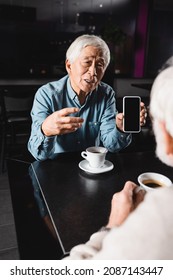 Senior Asian Man Pointing At Cellphone With Blank Screen While Spending Time With Blurred Friend In Cafe