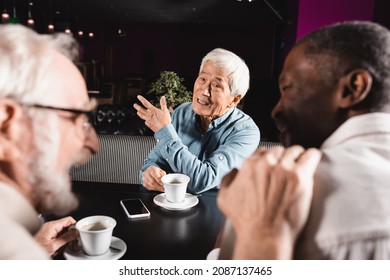 Senior Asian Man Pointing With Hand While Spending Time With Blurred Interracial Friends In Cafe