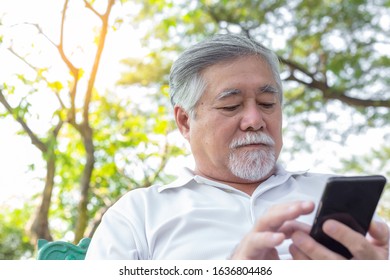 Senior Asian Man Reading News Or Checking Information On Website By Using Smart Phone And Internet Online At Park. Handsome Asian Elderly Man Has Nice Beard And Mustache. Old Guy Look Serious, Stress