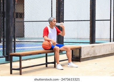 Senior Asian man in retirement age drinking water after playing basketball at outdoor court for healthy and fun activity to exercise and workout against aging illness - Powered by Shutterstock