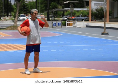 Senior Asian man in retirement age drinking water after playing basketball at outdoor court for healthy and fun activity to exercise and workout against aging illness - Powered by Shutterstock