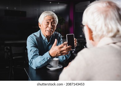 Senior Asian Man Showing Smartphone With Blank Screen To Blurred Friend In Cafe