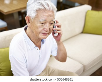 Senior Asian Man Sitting Alone On Sofa Talking On Mobile Phone Looking Sad.