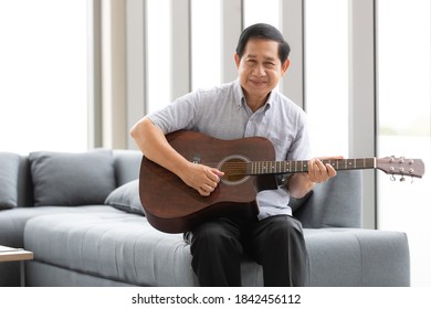 Senior Asian man sitting on sofa playing a guitar and looking to camera with a smile and kindly face. Idea for hobby and happiness free time of older people.