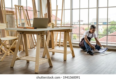 Senior Asian Man Sitting On Floor And Examining Draft While Working In Professional Carpenter Workshop. Home DIY In Free Time And Hobby Of Older Retired People.