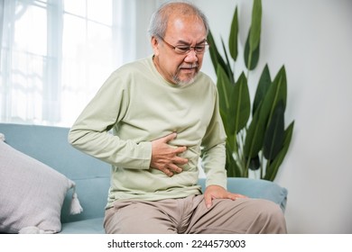 Senior Asian man sitting on sofa having suffering from stomach ache holding his stomach pain, Unhappy old aged man feeling discomfort because food poisoning, people health stomachache problem - Powered by Shutterstock