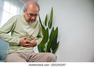 Senior Asian man sitting on sofa having suffering from stomach ache holding his stomach pain, Unhappy old aged man feeling discomfort because food poisoning, people health stomachache problem - Powered by Shutterstock