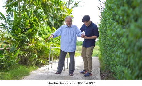 Senior Asian Man Struggling To Us A Walker. His Adult Son Is Helping Him