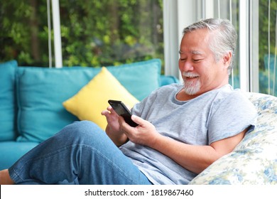 Senior Asian man using mobile phone while sitting in relaxation on the sofa couch in his pension cozy home